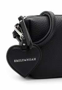 Schwarze Lederhandtasche mit strukturierter Oberfläche und herzförmigem Anhänger, der mit einem Metallclip befestigt ist. Der Anhänger ist mit den weißen Buchstaben "EMILY & NOAH" geprägt.