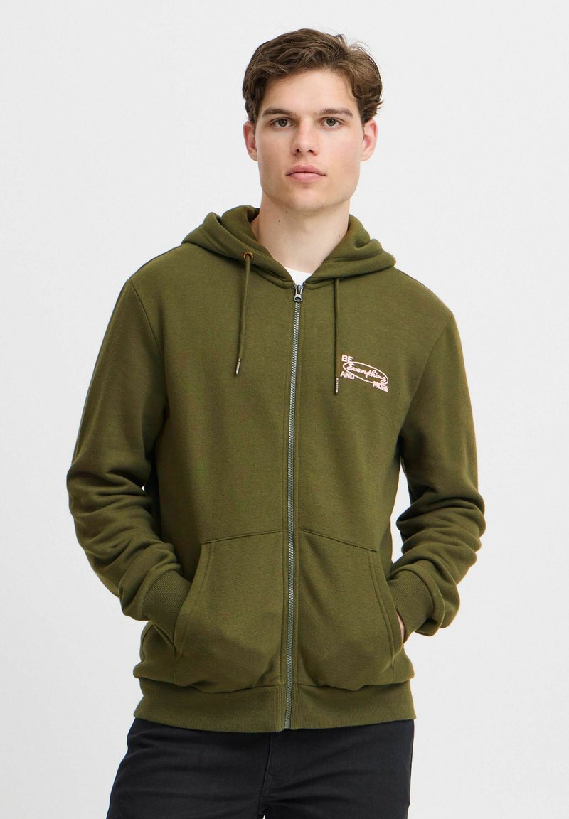 Olivenfarget zip-up hettegenser laget av mykt stoff, med trykk av logo foran, snorjustering i hetten og lommer foran. Enkel og funksjonell design.