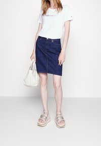 Bílé tričko s logem „HILFIGER“, tmavá denimová sukně tužkového střihu, béžové platformové sandály, nese světlou tašku přes rameno.