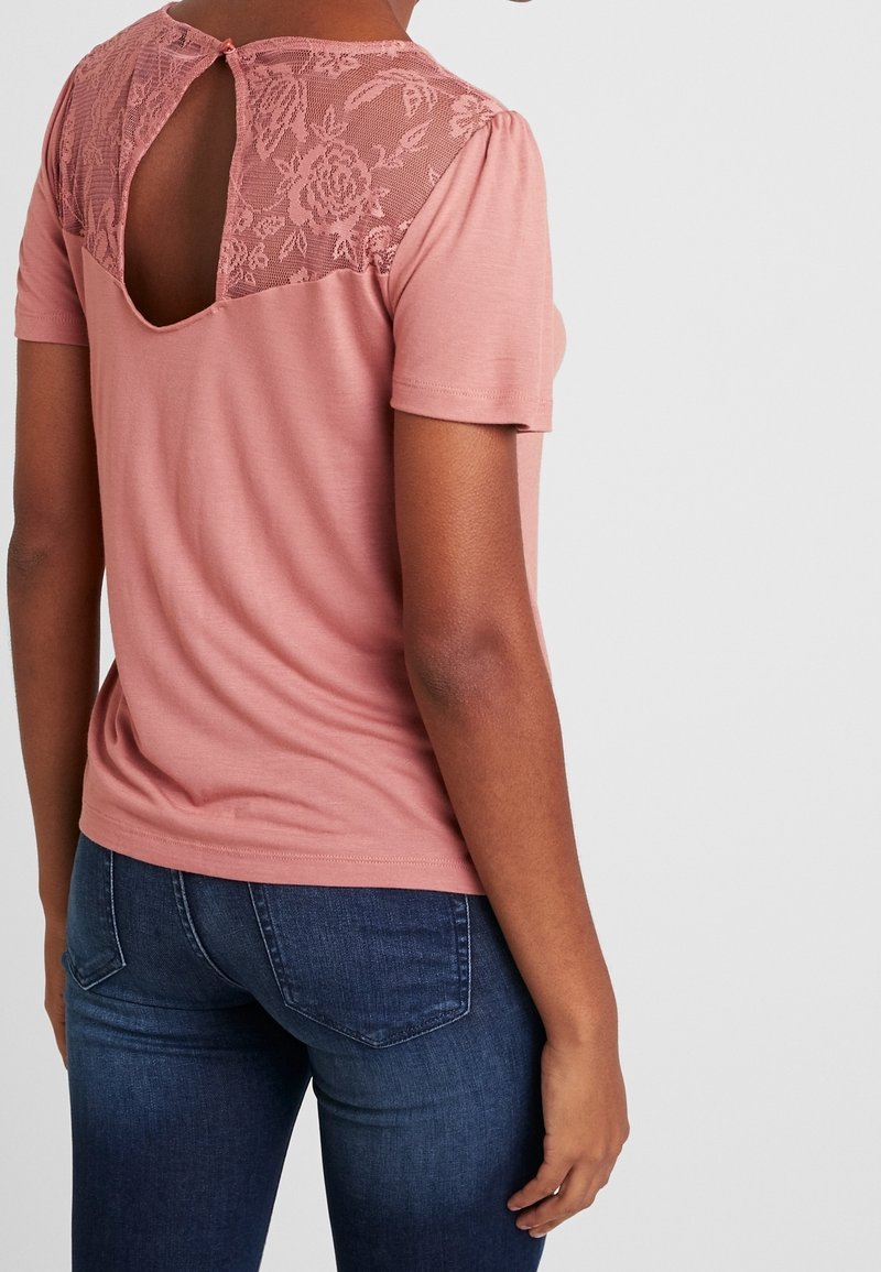 Lyserød kortærmet top med hængelukning i blondelook og nøglehuldetalje, blødt materiale og let rynkning ved ærmerne. Bæres med mørkeblå jeans.
