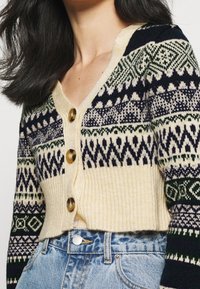 Gestrickte Strickjacke mit cremefarbener Basis, marineblauen und grünen geometrischen Mustern, V-Ausschnitt und vier großen braunen Knöpfen. Kurze, strukturierte Ärmel.