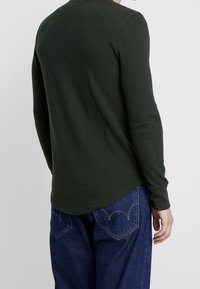 Camiseta de manga larga de color verde oscuro y textura acanalada, con cuello redondo, dobladillo curvado y mangas ajustadas, combinada con jeans azules que presentan costuras decorativas.