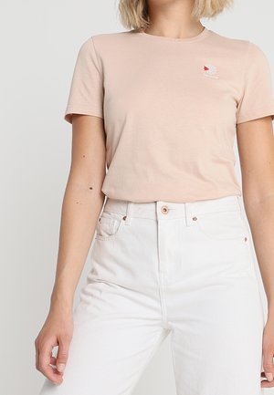 Kobieta ubrana w jasnoróżowy krótki rękaw t-shirt z małym logo i białe spodnie z wysokim stanem, stoi na jednolitym tle.