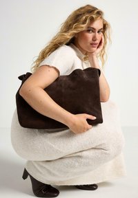 Borsa in suede marrone con una texture morbida e un bordo superiore ondulato. Modello che indossa un vestito a maglia color crema e stivali con tacco marroni.