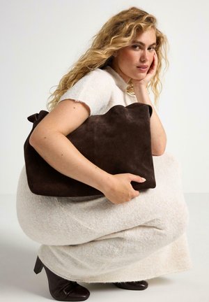 Borsa in suede marrone con una texture morbida e un bordo superiore ondulato. Modello che indossa un vestito a maglia color crema e stivali con tacco marroni.