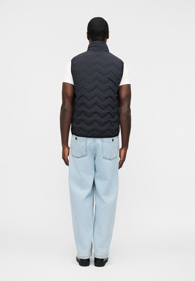 Άνδρας που φοράει μαύρο μπουφάν με καπιτονέ, λευκό κοντομάνικο πουκάμισο, ανοιχτό μπλε τζιν και μαύρα παπούτσια, στέκεται με την πλάτη προς το μέρος μας σε λευκό φόντο.