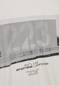 Biała koszulka z dużą szaro-skalową grafiką z napisem "223" oraz szczegółowym tekstem na dole. Design zawiera poziomy pas o teksturowanym wyglądzie.