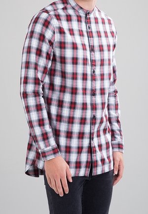 Homme portant une chemise à carreaux rouge, blanche et noire, boutonnée, avec un col mandarin et un pantalon sombre sur un fond uni.