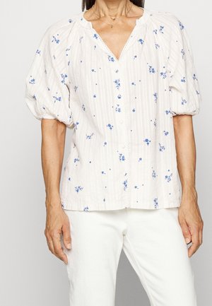 Kvinde iført hvid bluse med blå blomsterprint, puffede korte ærmer, knaplukning foran og hvide bukser.