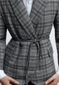 Grijze geruite blazer met een getailleerde silhoët, tailleband en twee voorzakken. De textuur van de stof heeft contrasterende zwart-witte patronen.