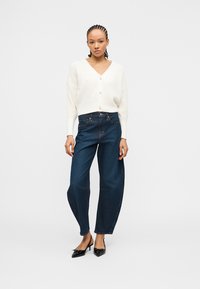 Femme debout portant un cardigan blanc boutonné, un jean taille haute ample bleu foncé, et des escarpins noirs à bout pointu avec des nœuds.