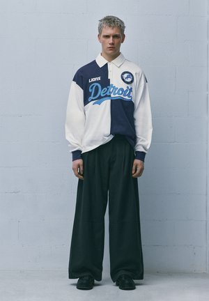 Jonge man met kort blond haar, gekleed in een tweekleurig Detroit Lions rugbyshirt en wijde zwarte broek, staand tegen een effen lichte muur.