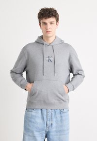 Pilkas megztinis su kapišonu, turintis kengūros kišenę ir Calvin Klein logotipą. Pagamintas iš minkštos medžiagos su raukšlėtomis rankovėmis ir apvadu.