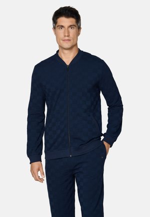 Mann trägt marineblaue Jacke mit Reißverschluss und passende Hosen mit dezentem Karomuster, steht mit einer Hand in der Tasche vor weißem Hintergrund.