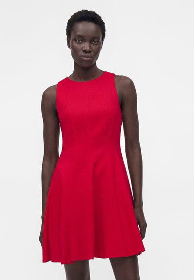 Γυναίκα με αμάνικο, φωτεινό κόκκινο, φόρεμα μέχρι το γόνατο με εφαρμοστή κορσέ και φουσκωτή φούστα μπροστά σε απλό ανοιχτό φόντο.