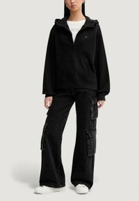 Černá mikina s kapucí na zip, s klokaní kapsou a logem. Kombinováno s černými cargo kalhotami a bílými teniskami.