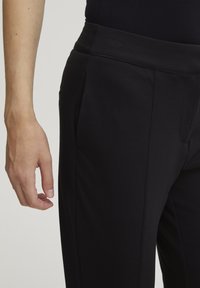Zwarte joggingbroek gemaakt van een gladde stof, met een brede tailleband, zijzakken en zorgvuldig gestikte naden, weergegeven met een hand die dichtbij de stof rust.