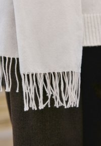 Hellbeiger Wollschal mit verdrehten Fransenenden, drapiert über dunklem strukturiertem Stoffhintergrund.