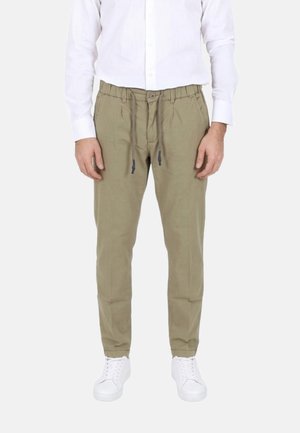 Uomo con pantaloni beige casual con coulisse, camicia bianca abbottonata e sneakers bianche, in piedi davanti a uno sfondo semplice.