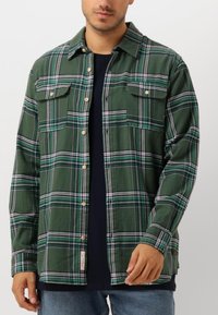 Camisa de franela a cuadros verdes con dos bolsillos frontales, botones de madera y corte relajado; presenta líneas blancas, grises y azules en el patrón.