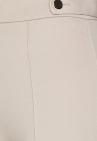 Beige høytlivs bukser med en glatt tekstur, med en svart knappdetalj og vertikale sømmer.
