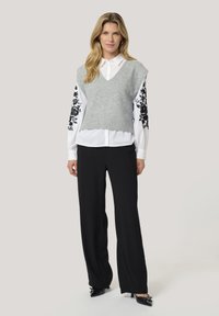 Woman wearing a white shirt with black floral embroidery on sleeves, a gray sleeveless sweater vest, black wide-leg pants, and black heels.