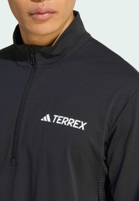 Black quarter-zip jacket with a high collar. Made of smooth fabric. Features a white "TERREX" logo and a zipper closure.