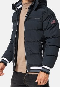 Tummansininen toppatakki, jossa huppu, valkoiset ja harmaat raidalliset hihansuut, vetoketjulliset taskut ja vasemmassa hihassa oleva merkki.