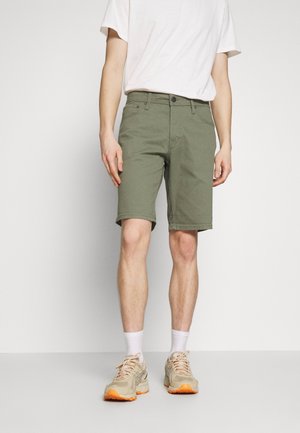 Pantalones cortos verde oliva con cuatro bolsillos, una cinturilla con botones y detalles de dobladillo cosidos. Llevado con una camiseta blanca básica y zapatillas beige.