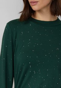 Zielona bluzka z długim rękawem i okrągłym dekoltem, zdobiona rozproszonymi srebrnymi ozdobami na miękkiej tkaninie. Prosty, nowoczesny fason.