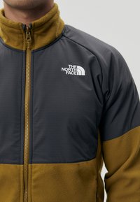 Giacca in pile con zip, caratterizzata da una parte superiore grigia e una parte inferiore gialla senape, con una finitura testurizzata e un logo bianco sul petto.