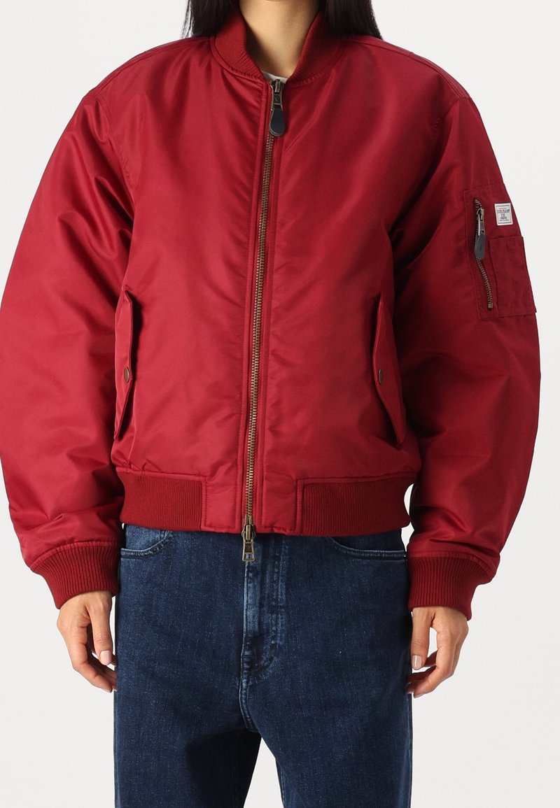 Personne portant une veste bomber rouge avec fermeture éclair à l'avant, poches latérales et une poche zippée sur la manche gauche, associée à un jean bleu.