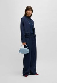 Marine bomberjack, bijpassende top en wijde broek. Lichtblauwe handtas met een gestructureerd ontwerp en subtiele hardware-details. Rode schoenen.