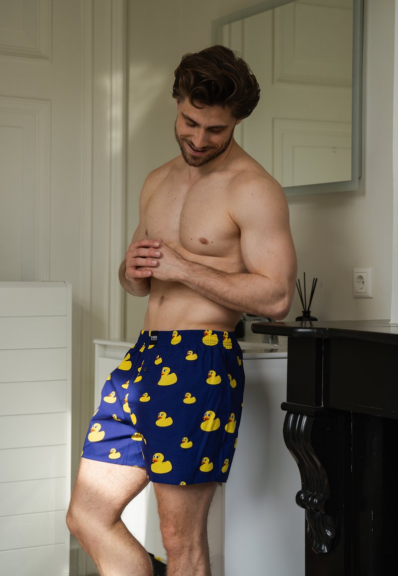 Blaue Badeshorts mit einem durchgehenden Muster aus gelben Gummi-Enten, einem elastischen Bund und einer entspannten Passform.