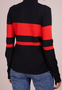 Schwarzer Pullover mit kräftigen roten horizontalen Streifen, ausgestattet mit einem hohen Kragen und gerippten Bündchen. Das Material scheint weich und ist in einer taillierten Silhouette gestaltet.