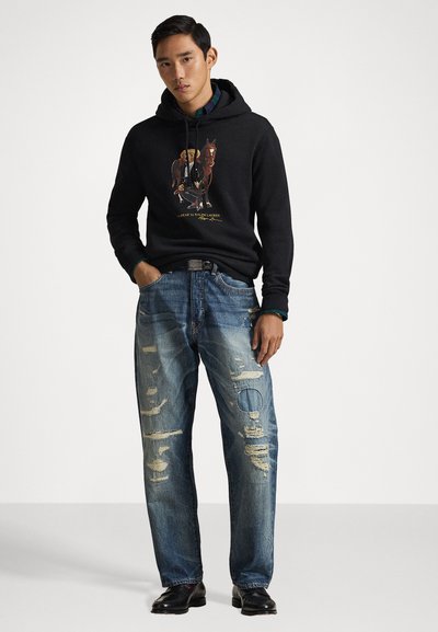 Juodas džemperis su grafiniu dizainu, derinamas su suplėšytais mėlynais džinsais, kuriuose yra dideli plyšiai ir apvalus skylė. Juodi batai užbaigia šį įvaizdį.