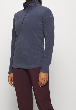 Personne portant un pull bleu marine à manches longues à fermeture éclair partielle et un pantalon bordeaux foncé sur un fond clair uni.