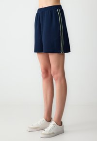 Pantaloni corti atletici blu navy con una vestibilità rilassata, caratterizzati da strisce verticali verdi e bianche sui lati. Realizzati in tessuto morbido e leggero.