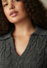 Pull en tricot gris avec un col en V, présentant un motif de câble texturé. La laine douce a une apparence confortable et épaisse.