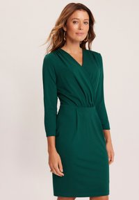 Zielona sukienka kopertowa z głębokim dekoltem w literę V, rękawami o długości 3/4 i dopasowanym taliowaniem. Wykonana z miękkiego materiału o gładkiej fakturze.