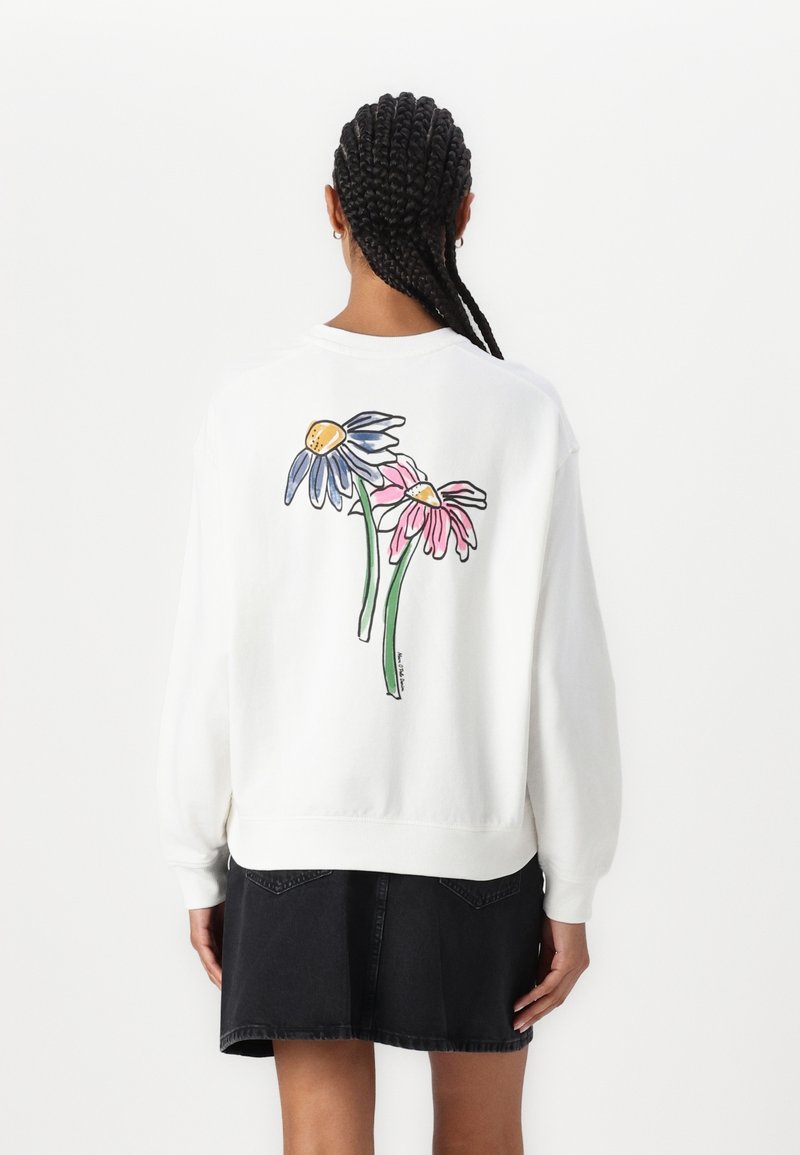 Femme aux cheveux tressés portant un sweat-shirt blanc avec des illustrations de fleurs bleues et roses dans le dos et une jupe en jean noire