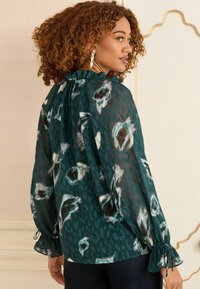 Blusa trasparente teal con stampa floreale e animale, scollatura arricciata e maniche gather. Tessuto leggero con motivi sottili.