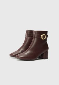 Bottines en cuir bordeaux avec un bout pointu, un talon carré et une accent en métal doré en forme de fleur sur le côté. Texture lisse.