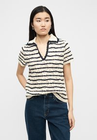 Νεαρή γυναίκα που φοράει εκρού και ναυτικό μπλε ριγέ πλεκτό τοπ με κοντά μανίκια και σκούρο μπλε τζιν, στέκεται με το ένα χέρι πίσω από την πλάτη σε λευκό φόντο.