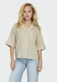Beige linned skjorte med korte Ærmer, en afslappet pasform, krave, brystlomme og hvide knapper. Kombineret med lyseblå jeans.