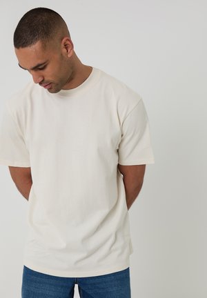 Homme aux cheveux courts portant une chemise à manches courtes uni blanc cassé et un jean bleu, regardant vers le bas avec les mains derrière le dos.