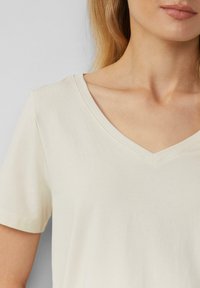 Kermanvärinen puuvillainen T-paita, jossa on V-pääntie ja lyhyet hihat. Paita on sileä ja siinä on vähäisesti ommeltuja yksityiskohtia pääntiellä.