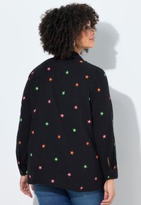 Vrouw met krullend haar die een zwart shirt met lange mouwen draagt, bedrukt met roze, groene en oranje sterren, zijwaarts gericht tegen een witte muur.