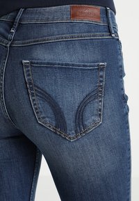 Jeans in denim blu scuro con un'etichetta in pelle marrone marchiata, dettagli della tasca posteriore con cuciture curve e una finitura liscia al tatto.