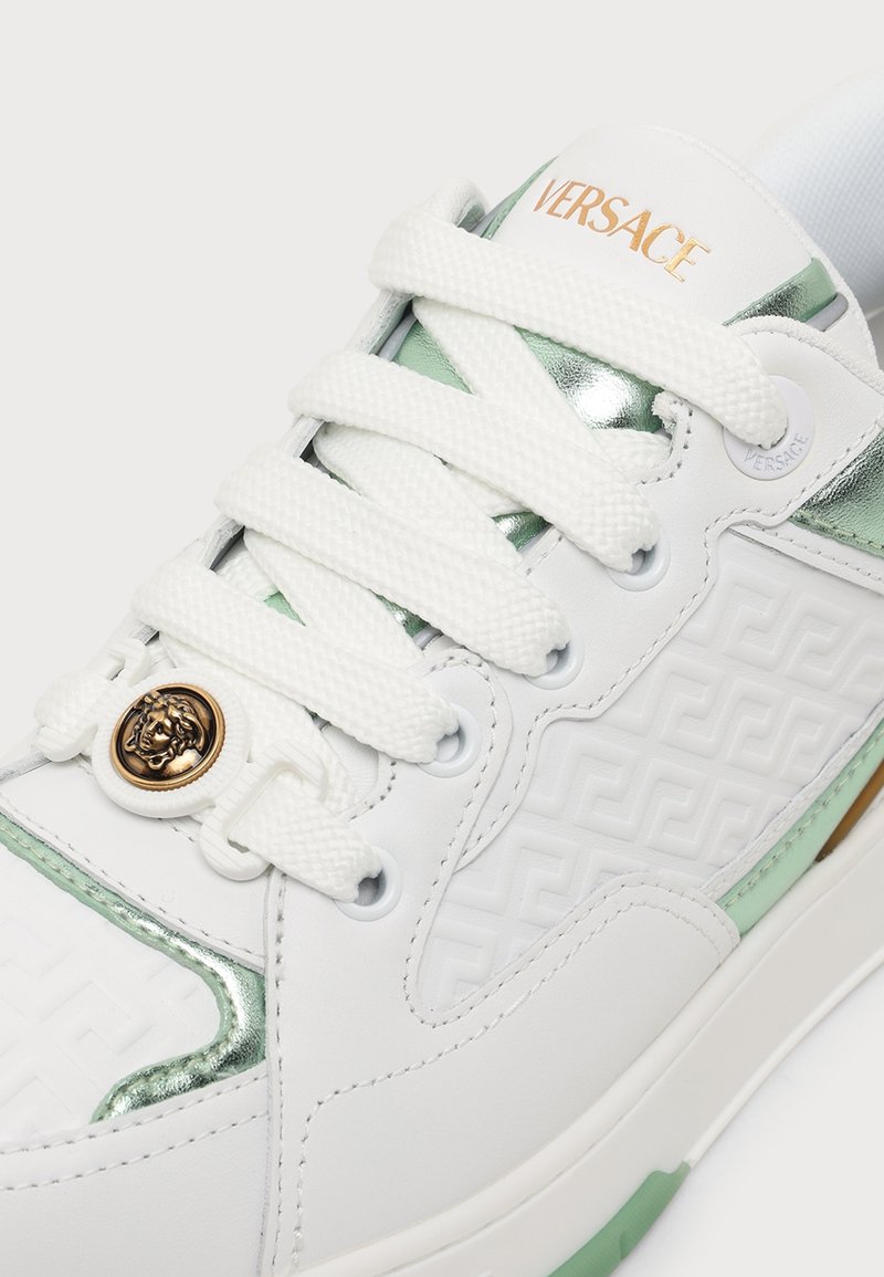 White Versace sneaker with gold logo on tongue, embossed geometric pattern, metallic green accents, and Medusa emblem on the laces.